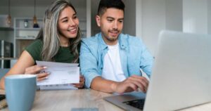 Happy Latin American couple at home paying bills online on their laptop and smiling
